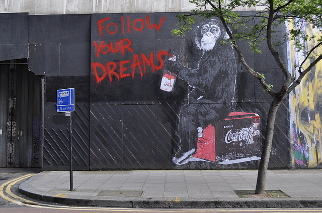 graffiti on a wall showing a monkey holding a can of red spray paint, with the words "follow your dreams"