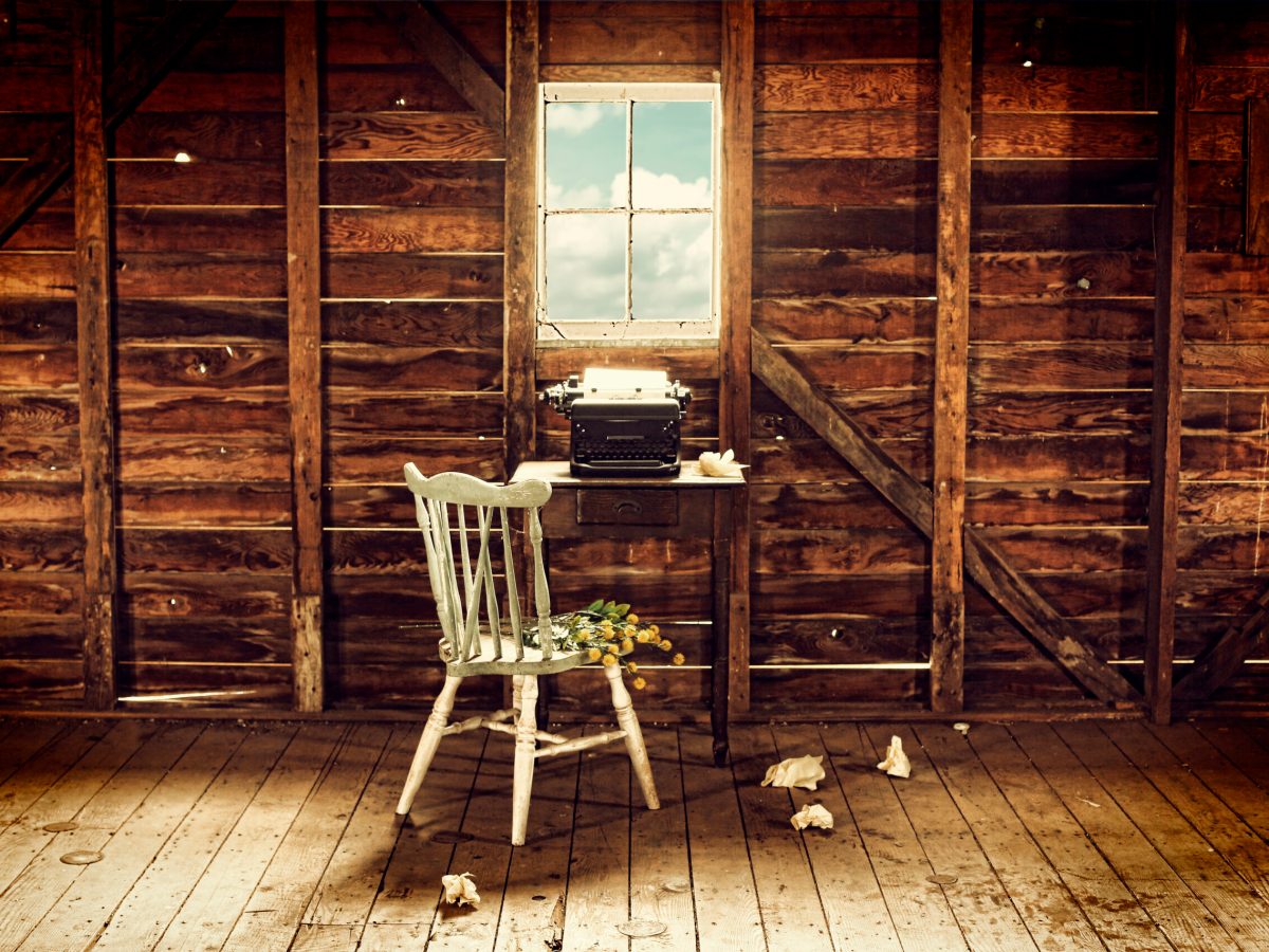 An empty chair sits in front of a typewriter and small desk in a desolate wood room with a view.