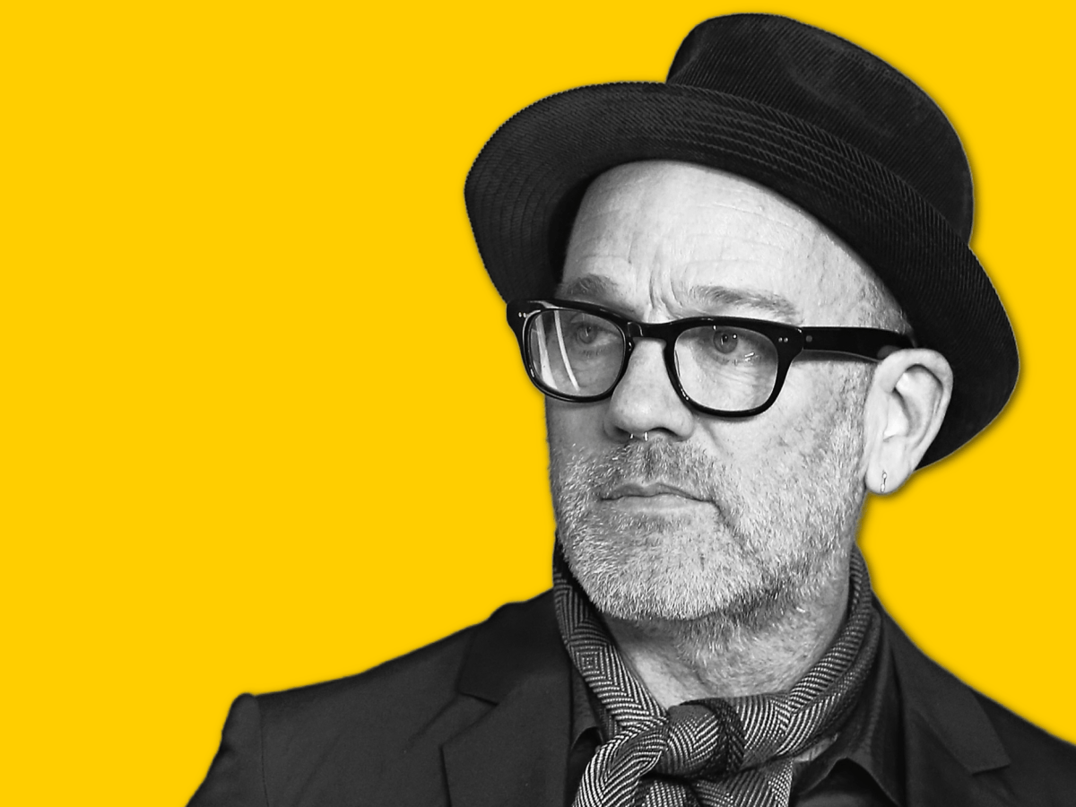 Singer Michael Stipe in a black hat and black glasses and suit against a yellow background